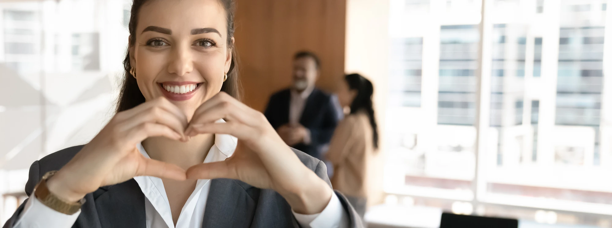 Smiling businesswoman making a heart shape with hands in the office, symbolizing teamwork and positivity.