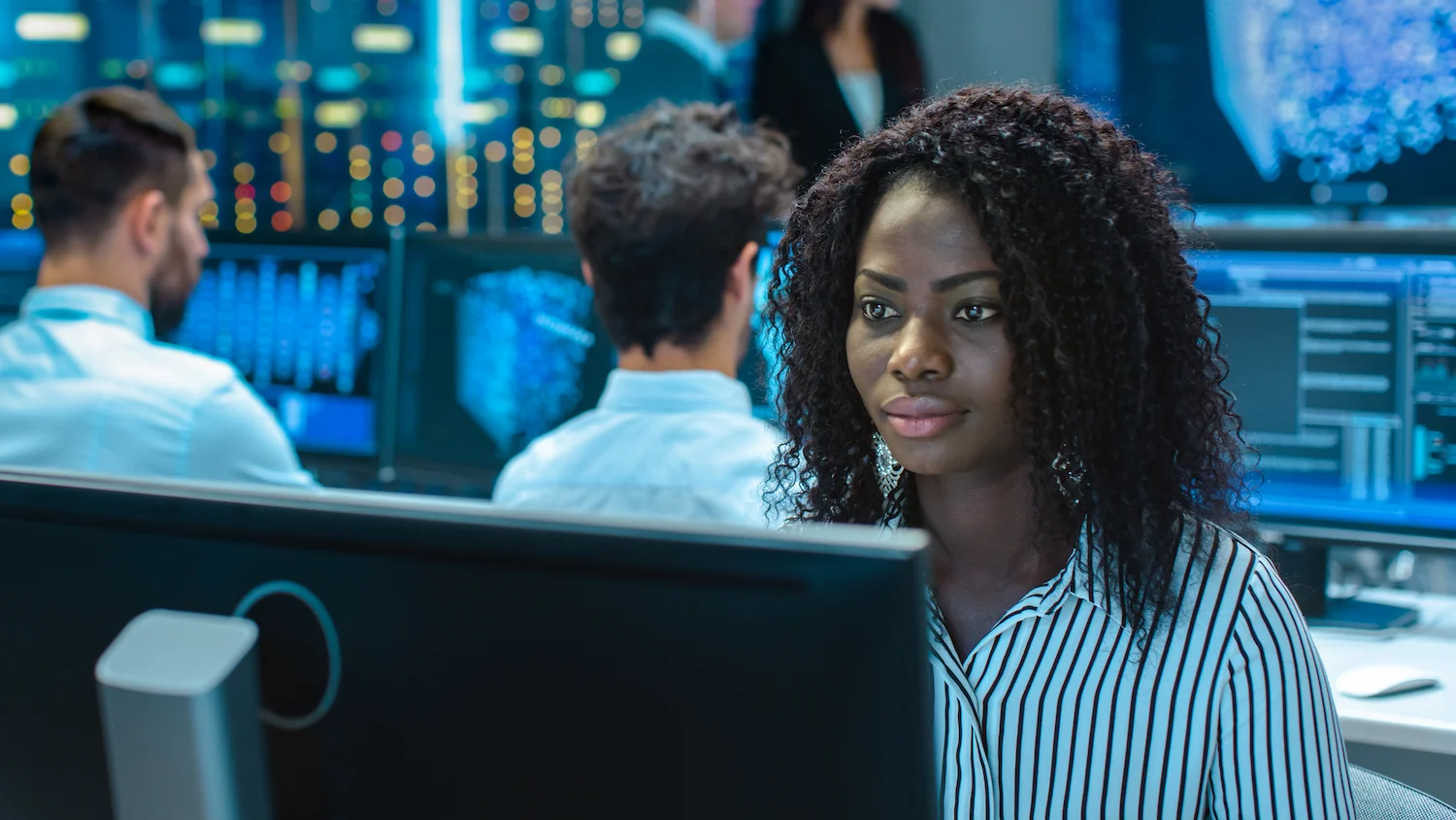 Woman working at a computer in a data center, focused on screen, colleagues and monitors visible in background.