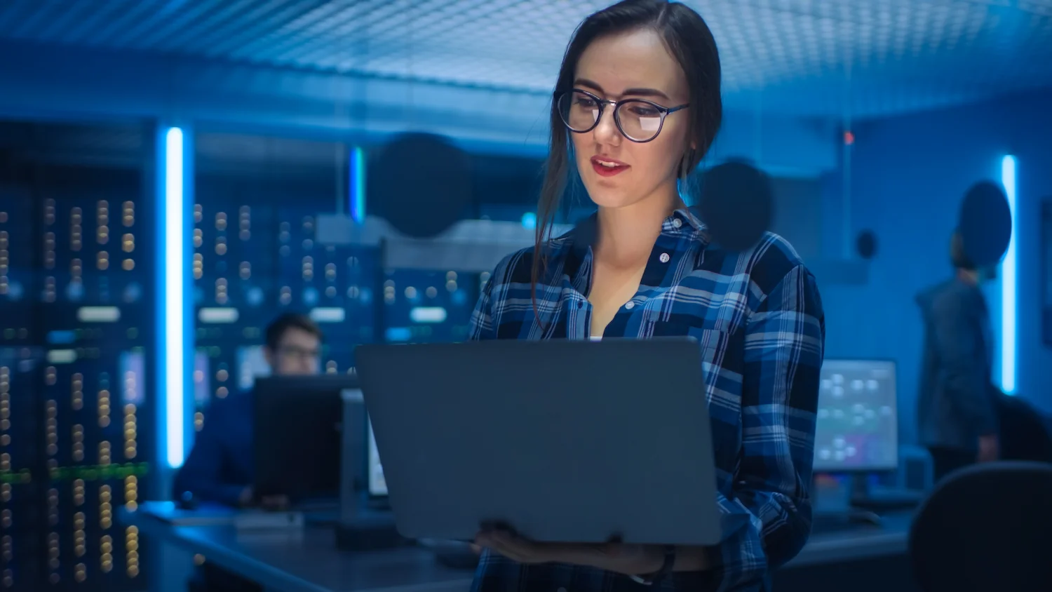 Woman in a data center working on a laptop, surrounded by screens and servers.