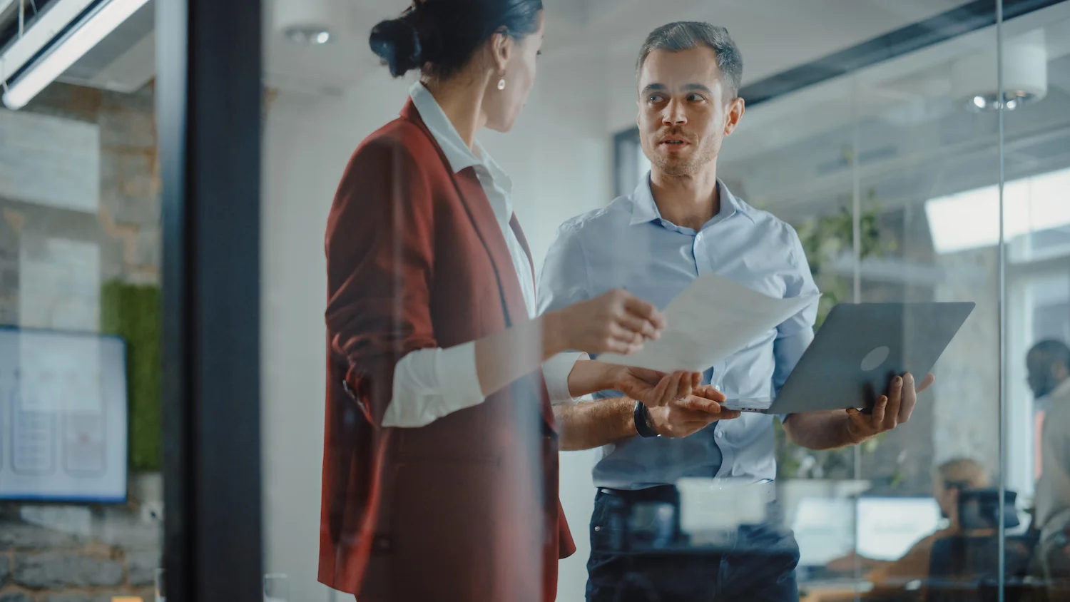 Business professionals in discussion, holding documents and laptop in modern office setting.