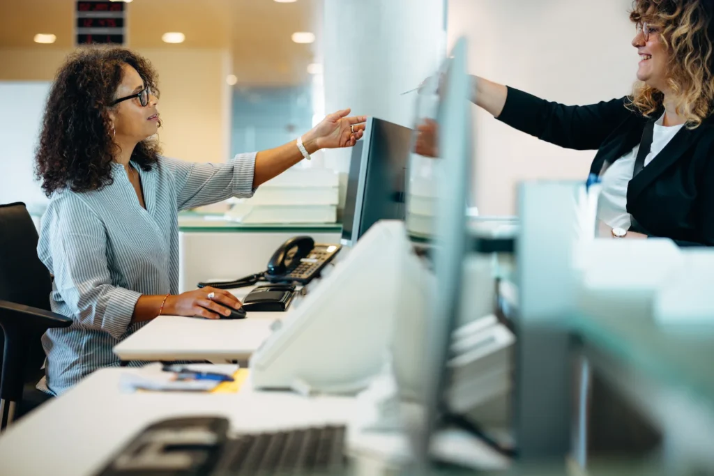 Two women working at a modern office desk, collaborating and exchanging documents between computer screens.