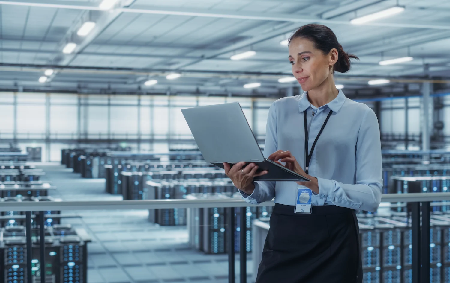 Woman using laptop in a large data center with rows of servers in the background, wearing business attire and a lanyard.