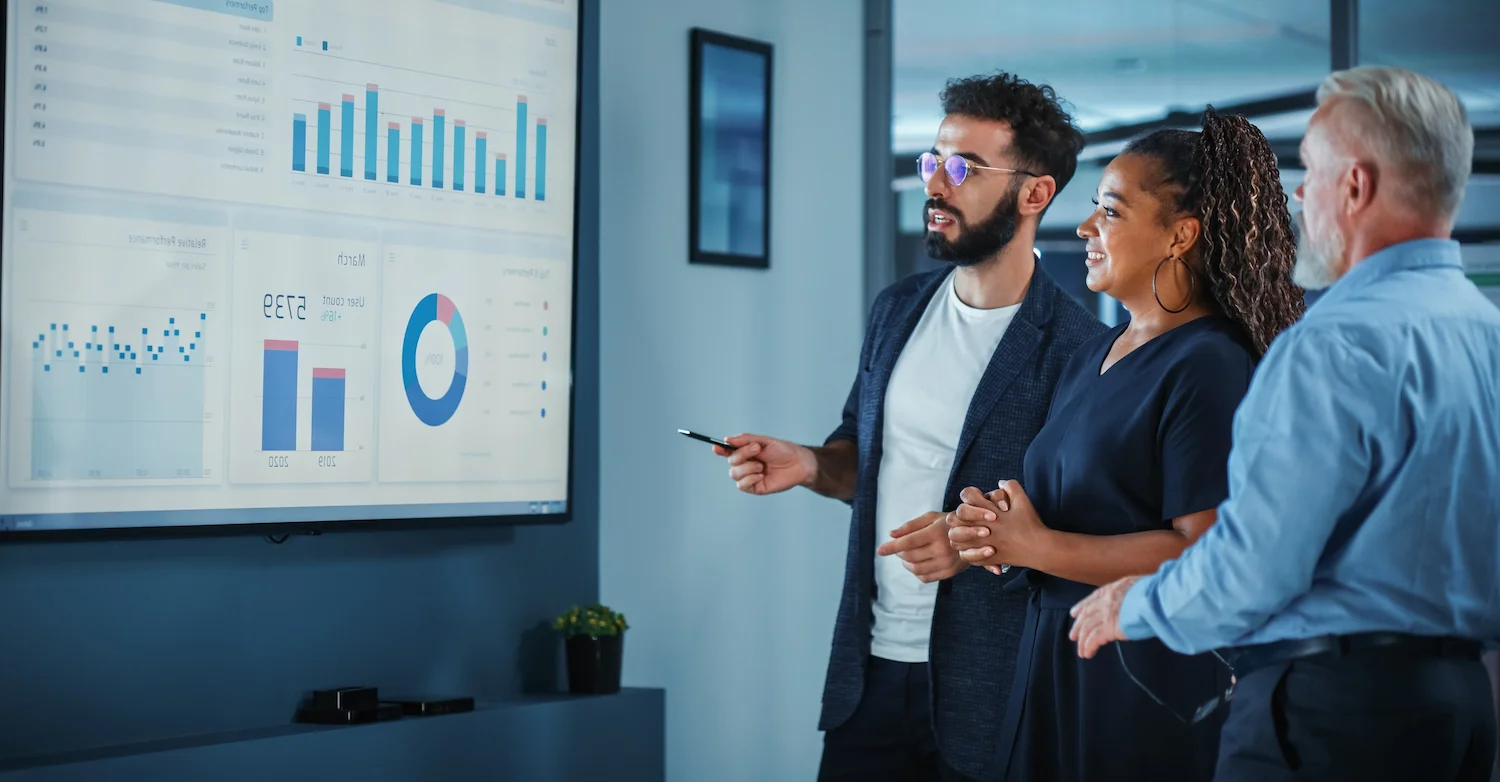 Team analyzing data on screen, discussing graphs and charts in modern office meeting.