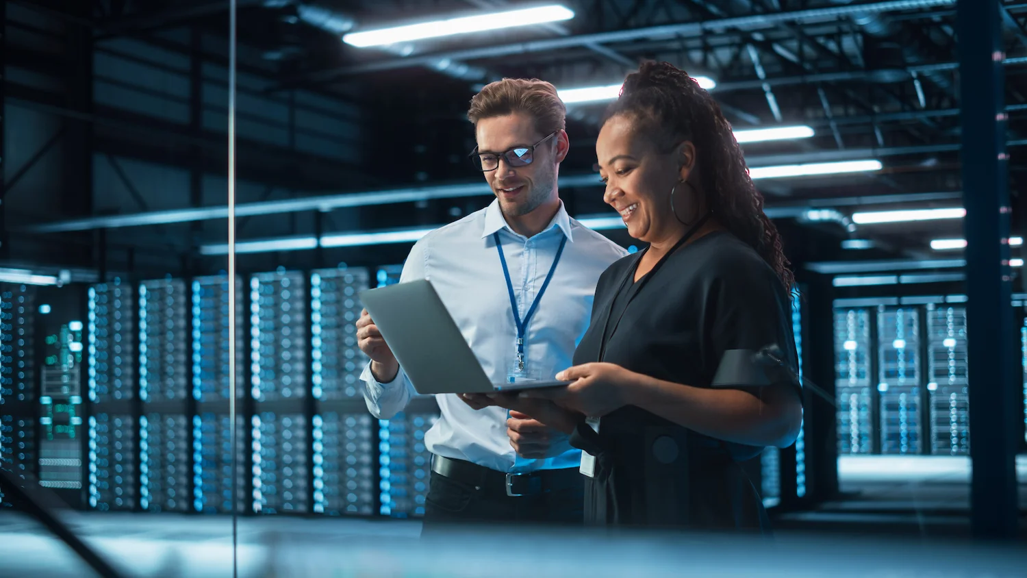 Two professionals smiling while using laptops in a data center server room.