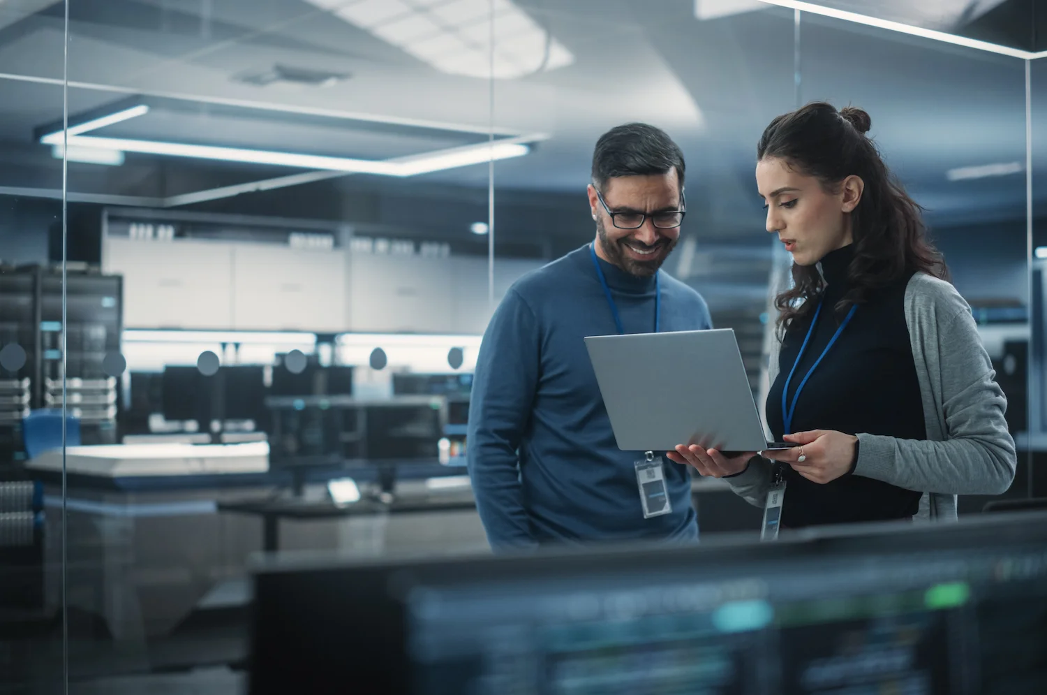 Two professionals collaborate on a laptop in a modern tech office with screens and servers in the background.