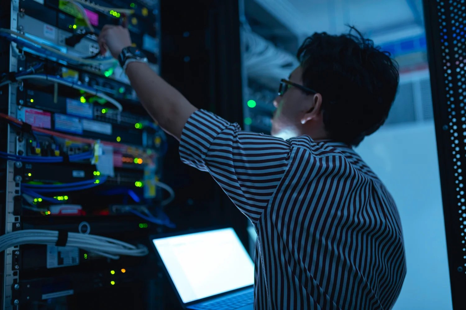 Technician managing servers in a data center, wearing a striped shirt and using a laptop.