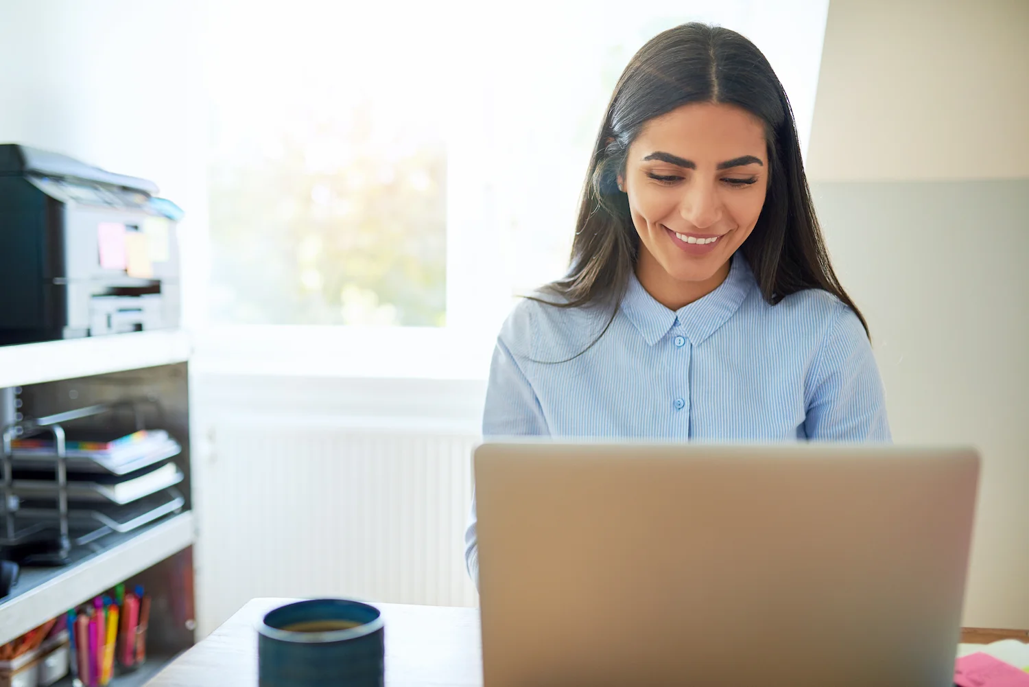 Smiling woman working on a laptop at home office desk, with bright window light.