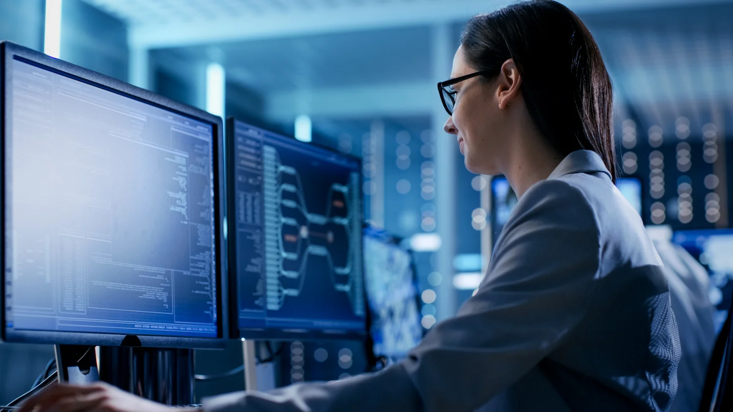Tech professional working at computer screens in a dark office, analyzing data and code on multiple monitors.