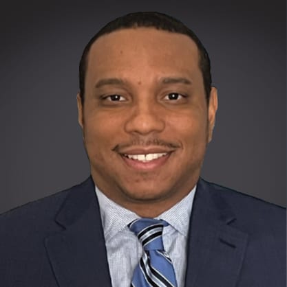 Professional headshot of a smiling man in a dark suit and striped tie, set against a dark background.