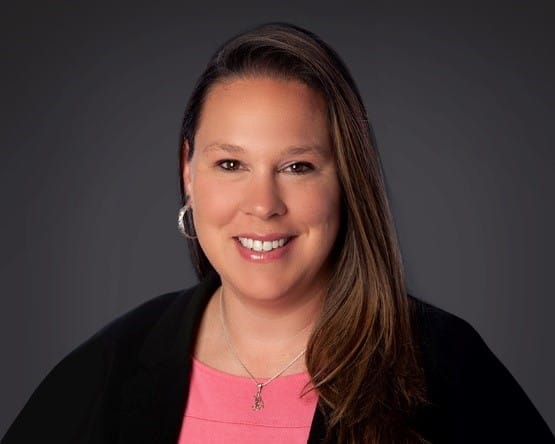 Professional headshot of a smiling woman with long brown hair, wearing a black blazer over a pink blouse, set against a dark gray background.