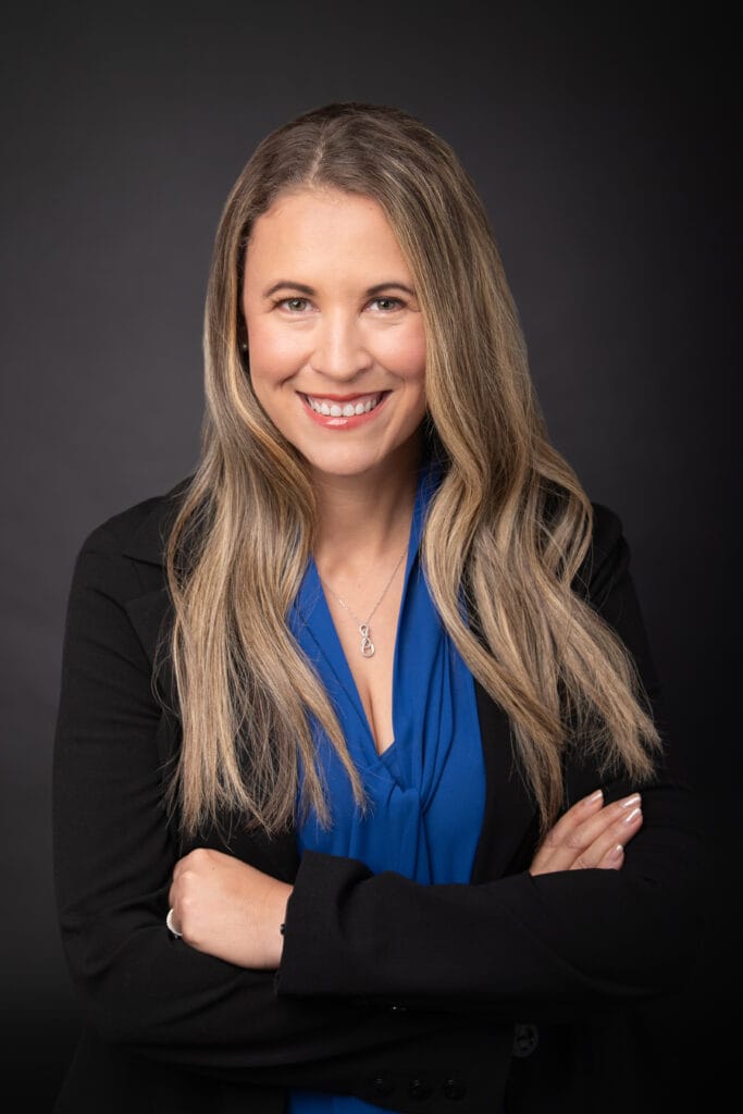 Professional headshot of a businesswoman in a dark suit and blue shirt, with a neutral expression and a blurred dark background, suitable for corporate profiles or business websites.