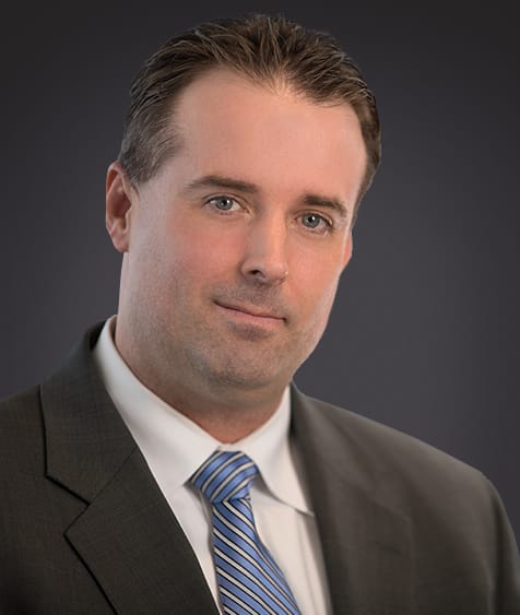Professional headshot of a businessman in a dark suit and blue tie, with a neutral expression and a blurred dark background, suitable for corporate profiles or business websites.