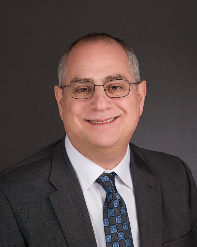 Professional headshot of a smiling man wearing glasses, a dark suit, white shirt, and a patterned tie, set against a neutral background. The image conveys a friendly and approachable demeanor suitable for corporate or business contexts.