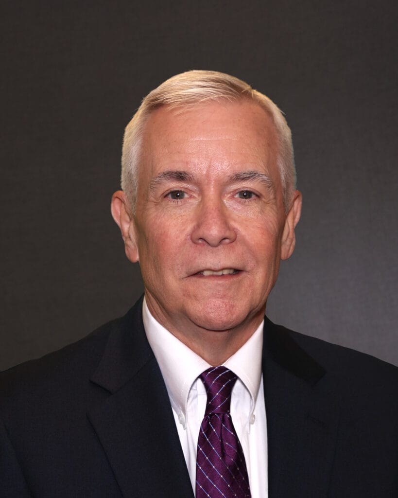 Professional headshot of a middle-aged man in a dark suit and a purple tie, with short gray hair and a neutral expression, set against a dark background.