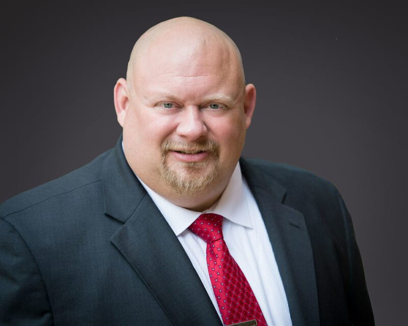Professional headshot of a bald man in a black suit and red tie, smiling against a dark background, suitable for business profiles or corporate websites.