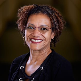 Smiling woman with curly hair and glasses, wearing a black top, against a blurred background.